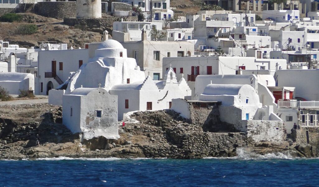 Whitewashed buildings and historic church clustered on the rocky coastline of Mykonos, viewed from the Aegean Sea.