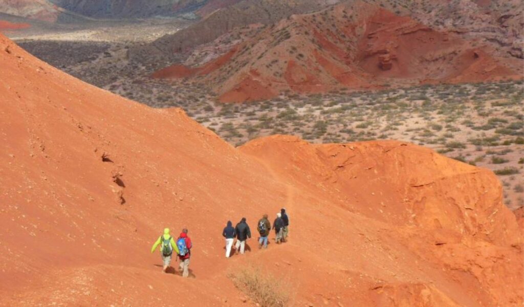 Backpackers hike along a bright red desert trail with colorful mountains in the distance under a cloudy sky.