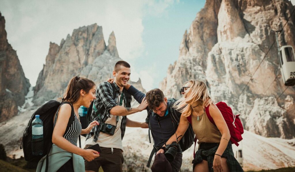 Group of happy hikers laughing together in a mountain valley with backpacks and cameras, surrounded by towering rocky peaks and a cable car in the background.