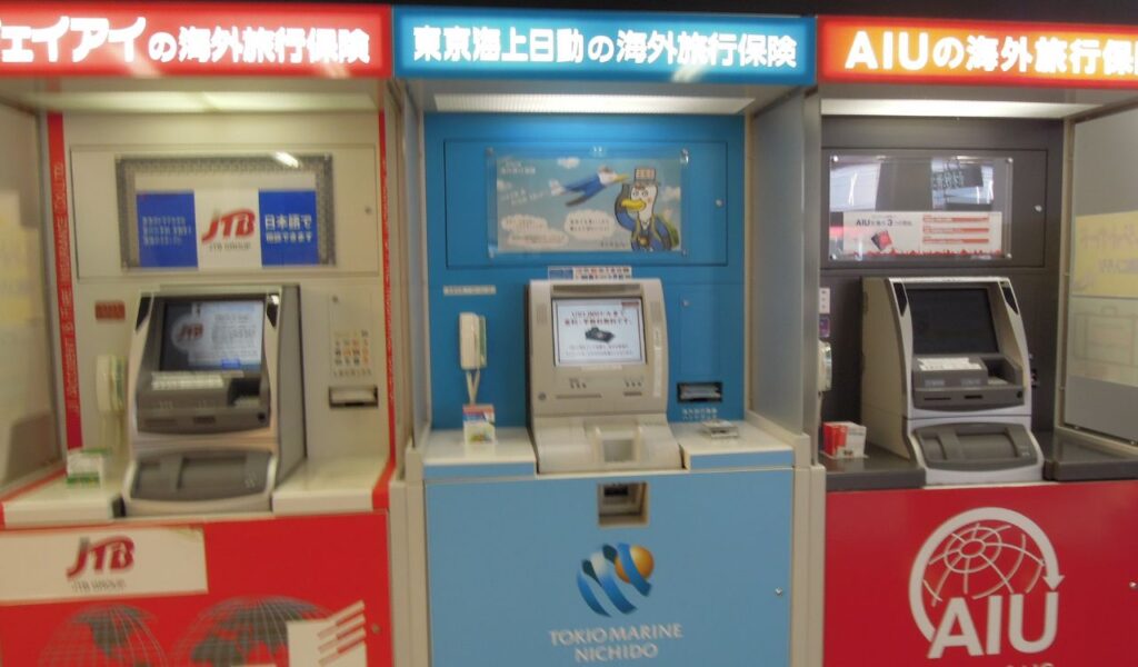 Row of red and blue Japanese travel‑insurance kiosks at an airport, displaying company logos and small touch‑screen terminals.