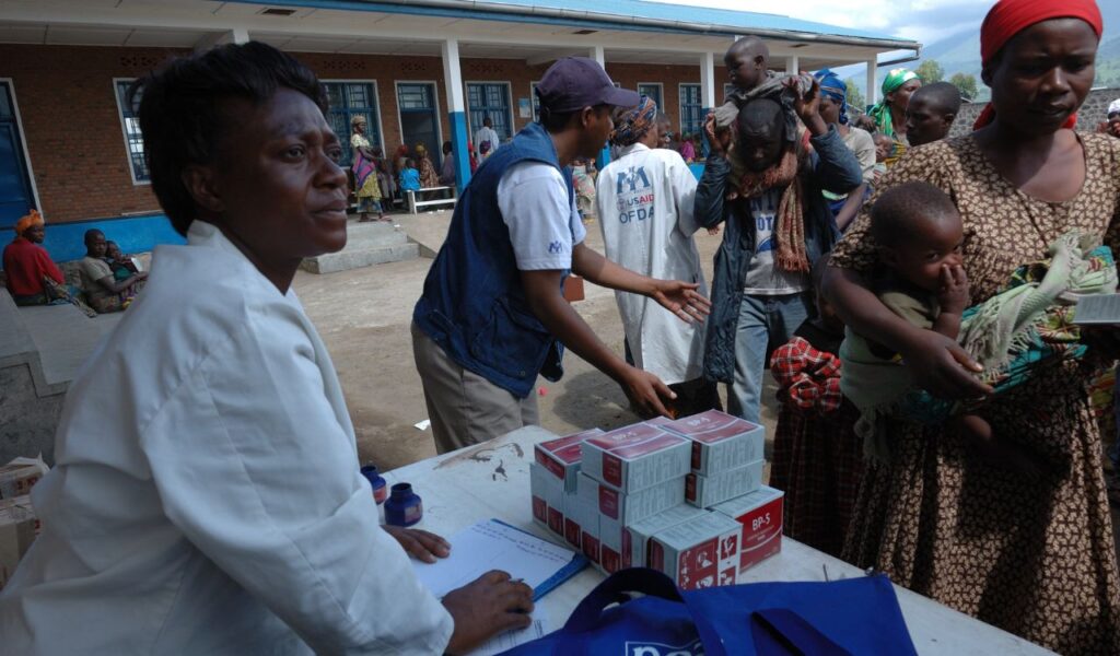 Aid worker hands out boxed medical supplies to worried mothers and children gathered outside a rural clinic.