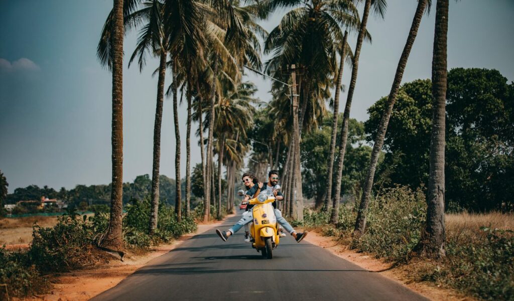 A joyful couple rides a yellow scooter down a palm-lined road on a sunny day, evoking a carefree vacation vibe.