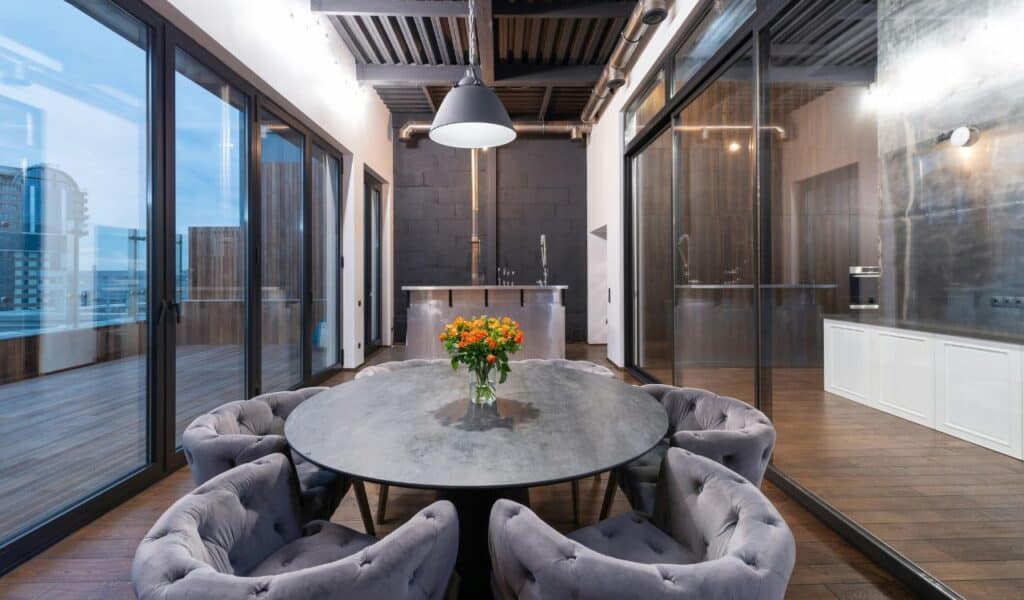 Modern dining area with a round stone table, tufted velvet chairs, and a vase of orange flowers, surrounded by glass walls and an industrial-style ceiling.