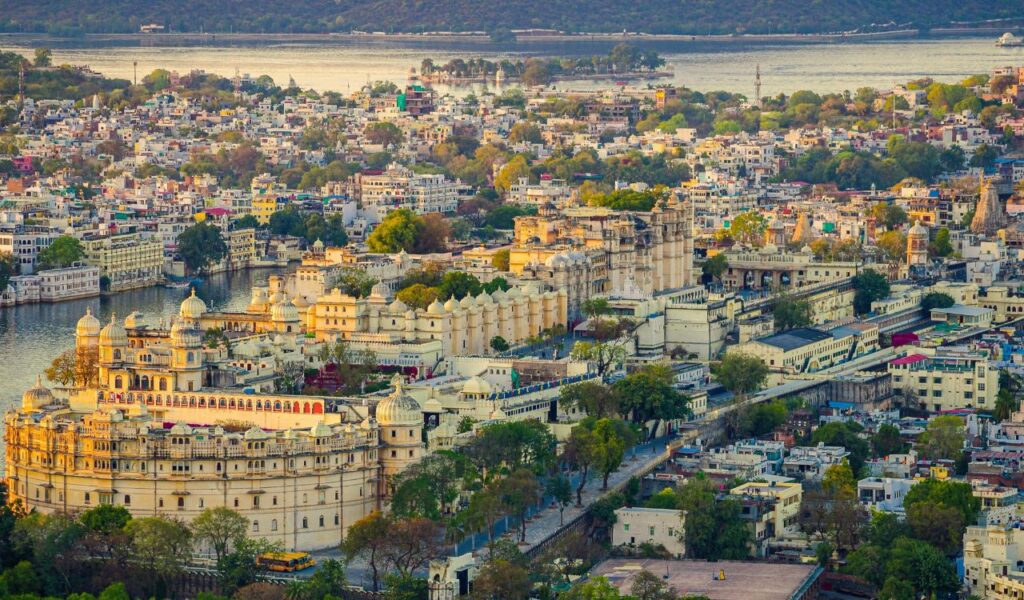 Aerial view of Udaipur, India, highlighting the grand City Palace complex beside Lake Pichola with surrounding historic architecture and distant hills.