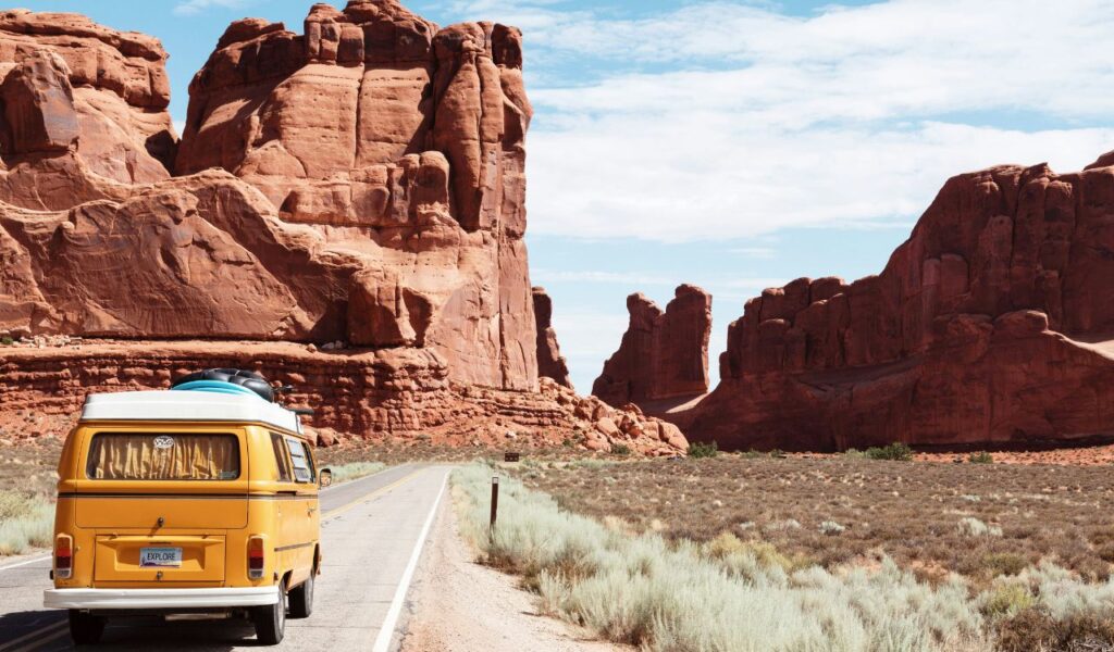 A yellow van with a surfboard on top drives through a desert road flanked by massive red rock formations under blue skies.