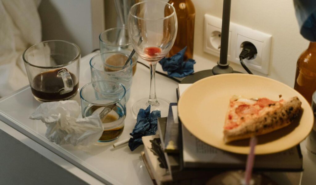 Messy nightstand with empty glasses, crumpled napkins, books, and a plate holding a single slice of pepperoni pizza.
