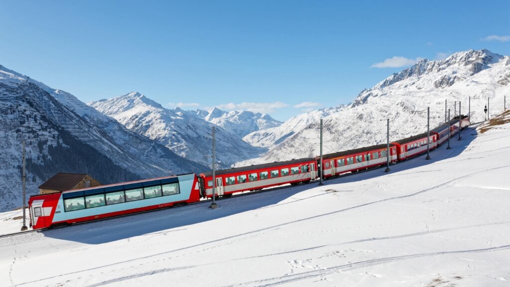 Tourists ride on a Glacier Express of Matterhorn Gotthard Railway on a sunny winter day, enjoying the view thru panoramic windows of snowy mountains under blue clear sky in Andermatt, Uri, Switzerland