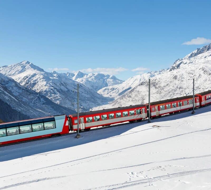 Tourists ride on a Glacier Express of Matterhorn Gotthard Railway on a sunny winter day, enjoying the view thru panoramic windows of snowy mountains under blue clear sky in Andermatt, Uri, Switzerland