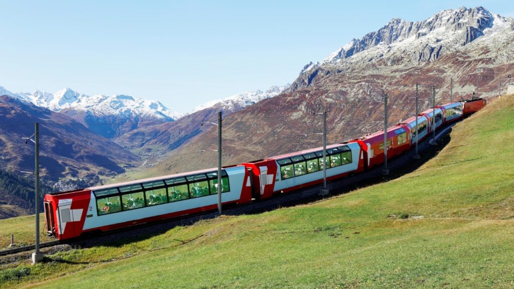 Tourists ride a Glacier Express on Matterhorn Gotthard Railway on a brisk fall day, enjoying the view thru panoramic windows of the snowy mountains under blue clear sky, in Andermatt, Uri, Switzerland