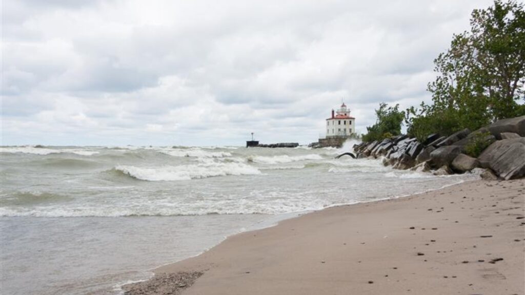 Lighthouse at Mentor Headlands Beach State Park in Ohio with waves crashing on shore and dark clouds overhead.