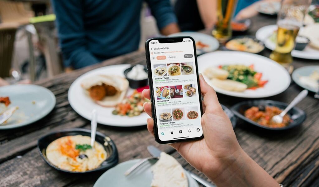 A person holding a smartphone with a restaurant review app open, displaying local food options, while sitting at a table filled with various dishes and drinks during an outdoor meal