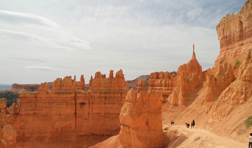 Hikers walk along a dusty trail beneath towering orange hoodoos in Bryce Canyon National Park under a partly cloudy sky.