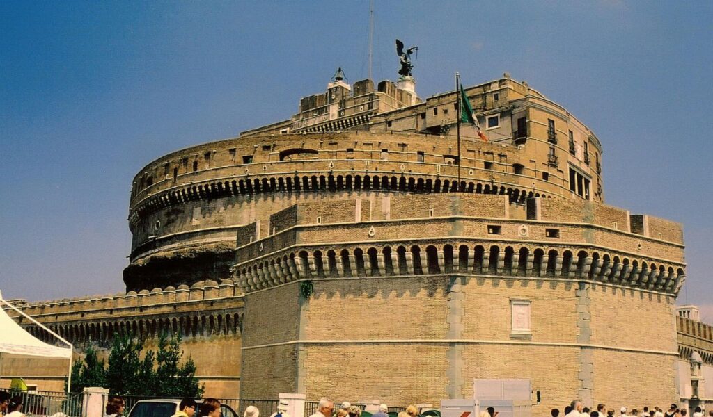 Exterior view of Castel Sant'Angelo in Rome, with its circular fortress structure, Italian flag, and crowds of visitors on a sunny day.