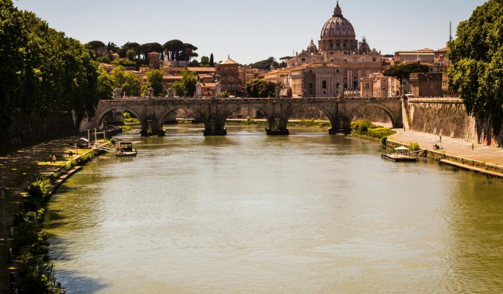 Scenic view of Vatican Gardens with the Tiber River, Ponte Sant'Angelo bridge, and St. Peter's Basilica in the background on a sunny day.