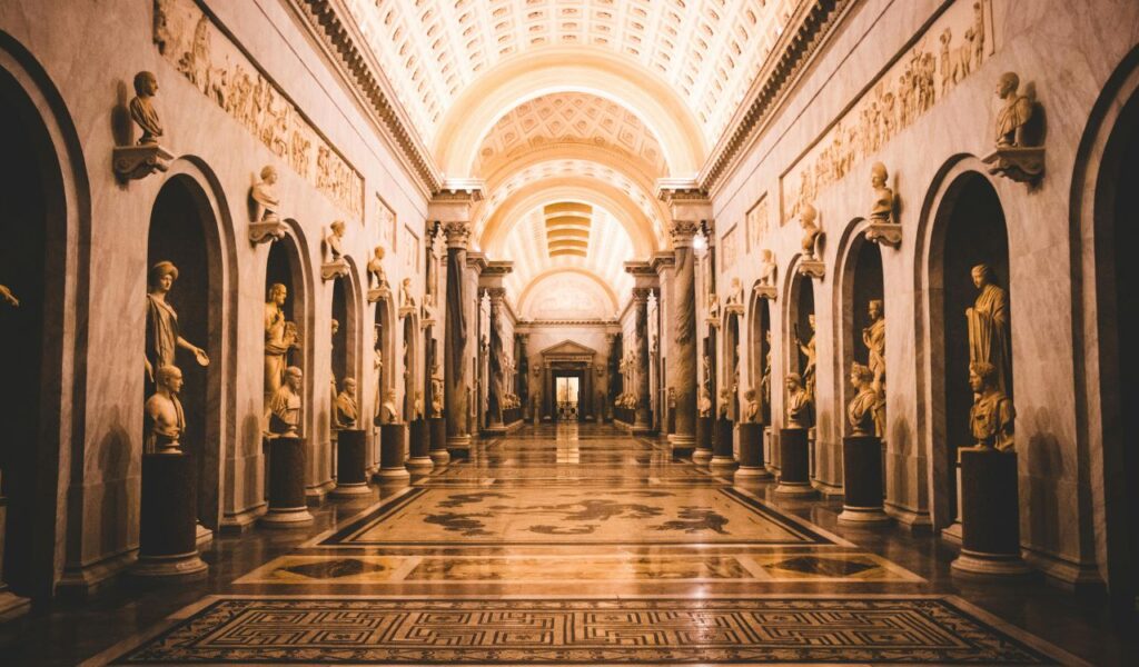 rnate hallway inside the Vatican Museums featuring classical statues, arched niches, intricate floor patterns, and a glowing vaulted ceiling.