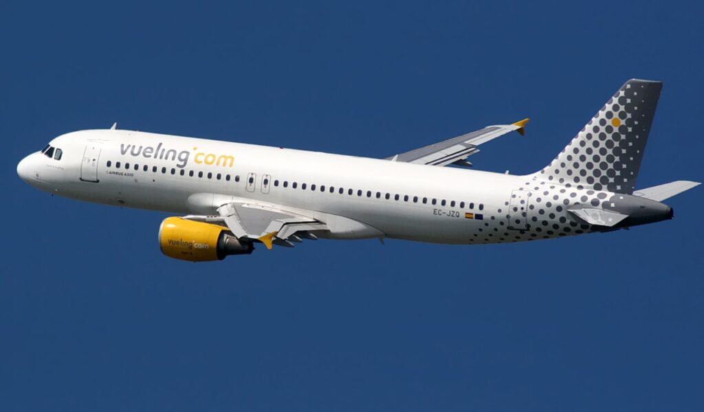 Vueling Airlines jet in flight, featuring a clean white fuselage, yellow engines, and dotted grey tail design.