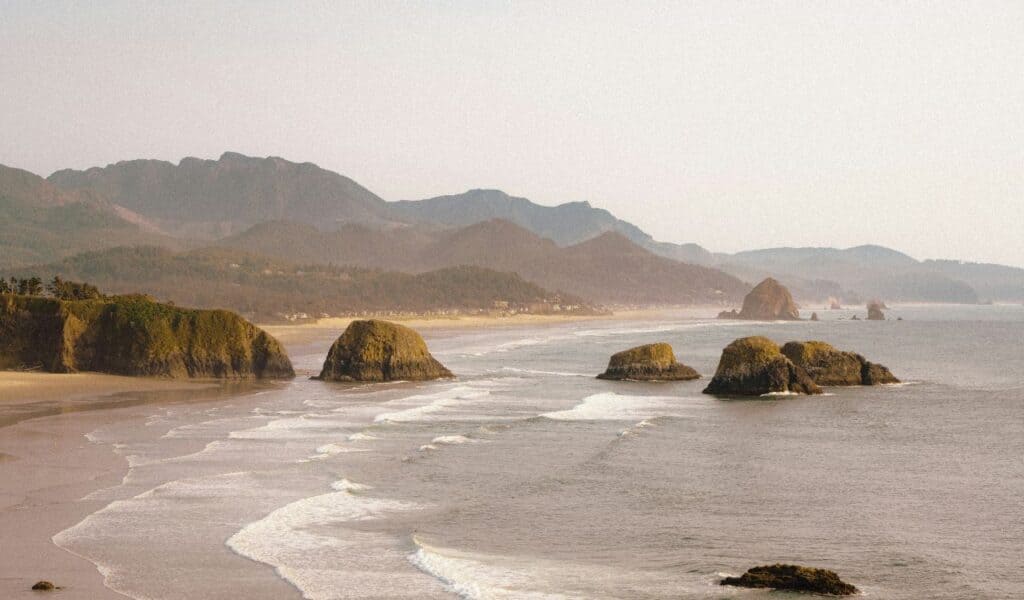 Rocky sea stacks along the shore at Battle Rock Wayside Park with waves and forested hills in the background.