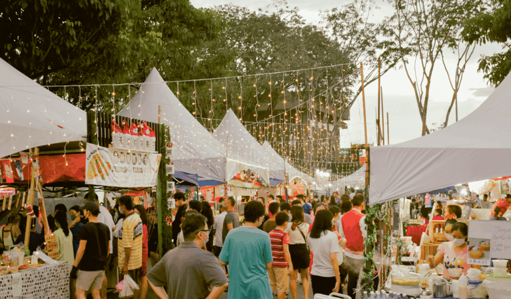 A lively evening market scene with rows of white tents, hanging string lights, and crowds of people exploring food stalls and local vendors under the trees.