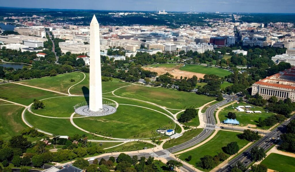 Aerial view of the Washington Monument surrounded by the National Mall, green lawns, pathways, and city buildings in Washington, D.C.