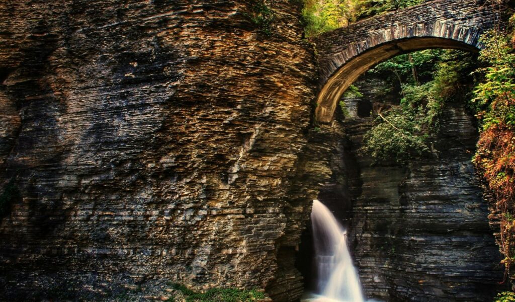 A narrow waterfall flows through a rocky gorge beneath a stone bridge surrounded by layered cliffs and greenery.