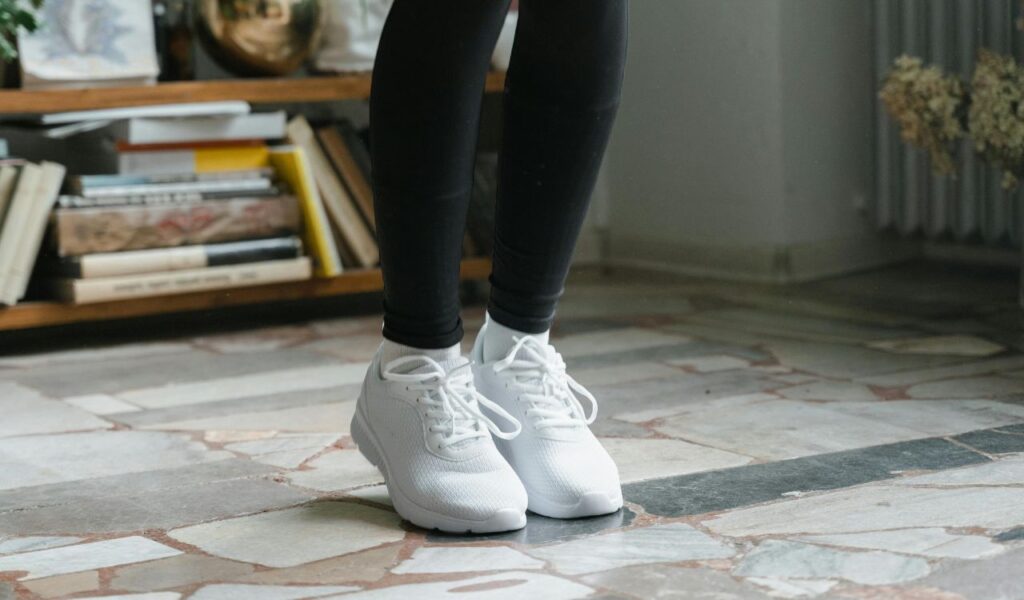 Close-up of a person wearing white sneakers and black leggings standing indoors on a tiled floor with bookshelves in the background.