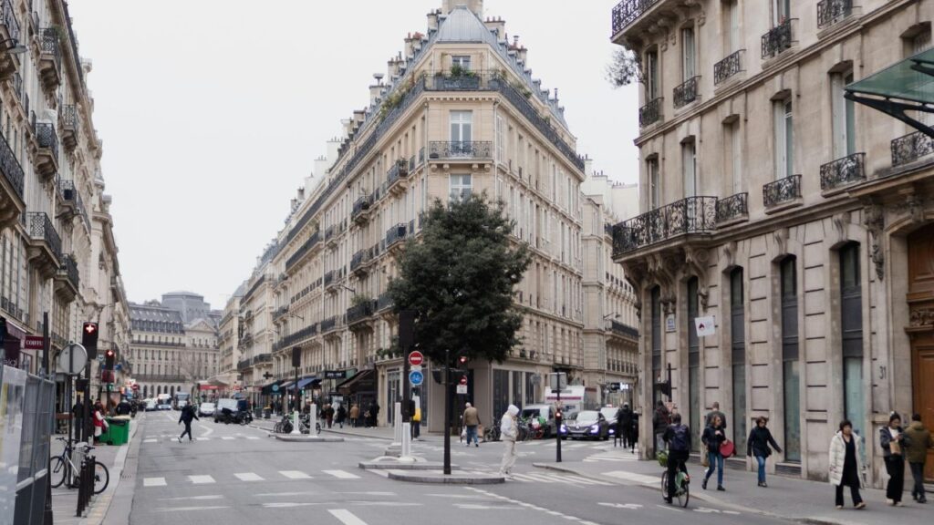 A bustling Paris street scene with classic Haussmann-style buildings, pedestrians walking, cyclists riding, and cars navigating the intersection.