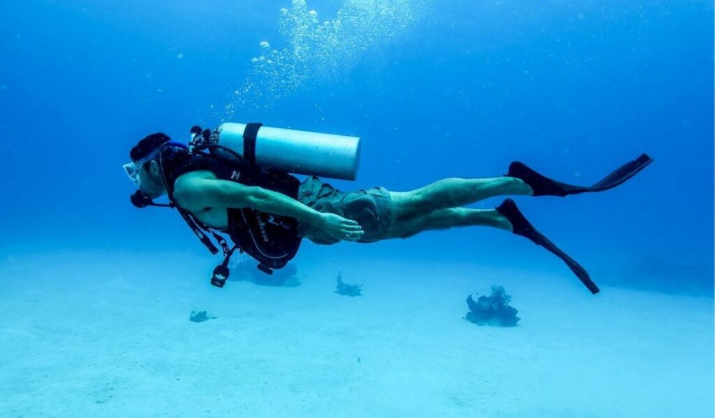 Scuba diver swimming underwater near the ocean floor, wearing fins and a tank, representing the need for beach safety awareness.