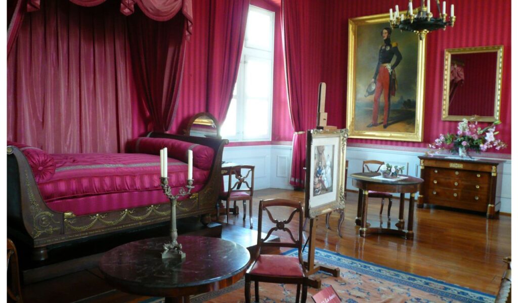 A lavish room with bold red drapes, satin upholstery, ornate furniture, and mismatched classical decor fills the eclectic space.