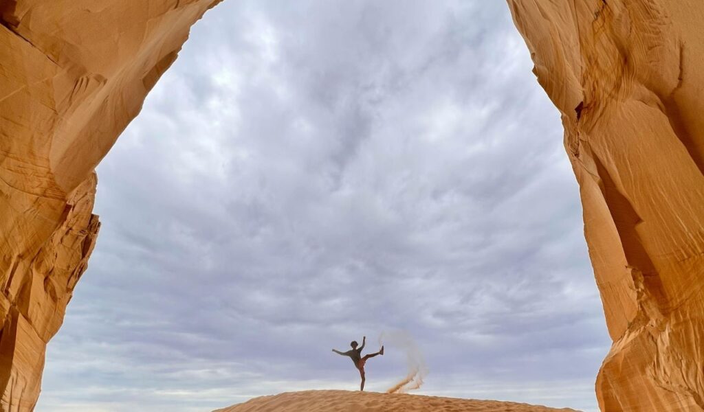A person kicks up sand while posing beneath towering red rock walls, framed by a natural arch under a dramatic cloudy sky.