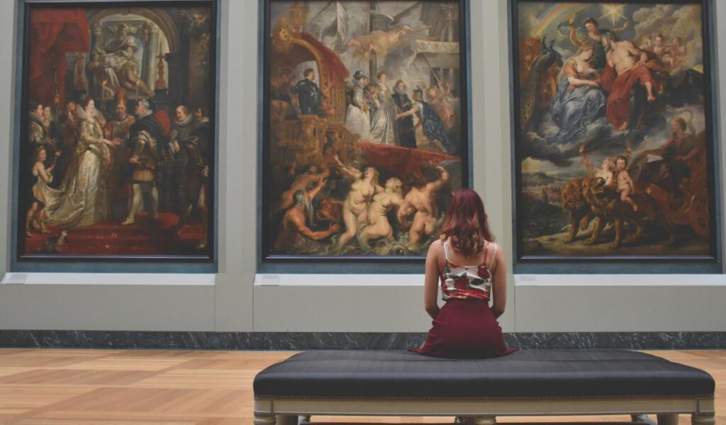 Woman sitting on a bench in an art museum, admiring large classical paintings displayed on the gallery wall.