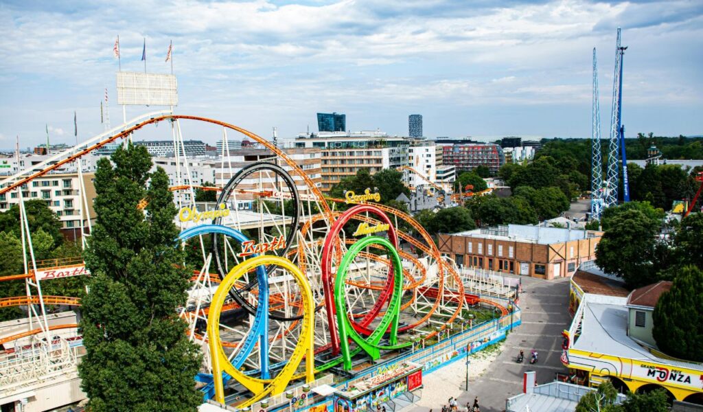 Colorful roller coaster with multiple loops at Wurstelprater amusement park in Vienna, Austria, surrounded by trees and urban buildings.