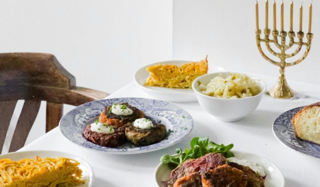 A table set with Yerushalmi kugel, latkes topped with sour cream, cooked cabbage, bread, and a lit menorah in the background.