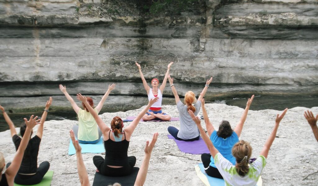 A yoga instructor leads a group of people in an outdoor yoga session on rocky terrain near a cliffside, all seated on colorful mats with arms raised in unison.