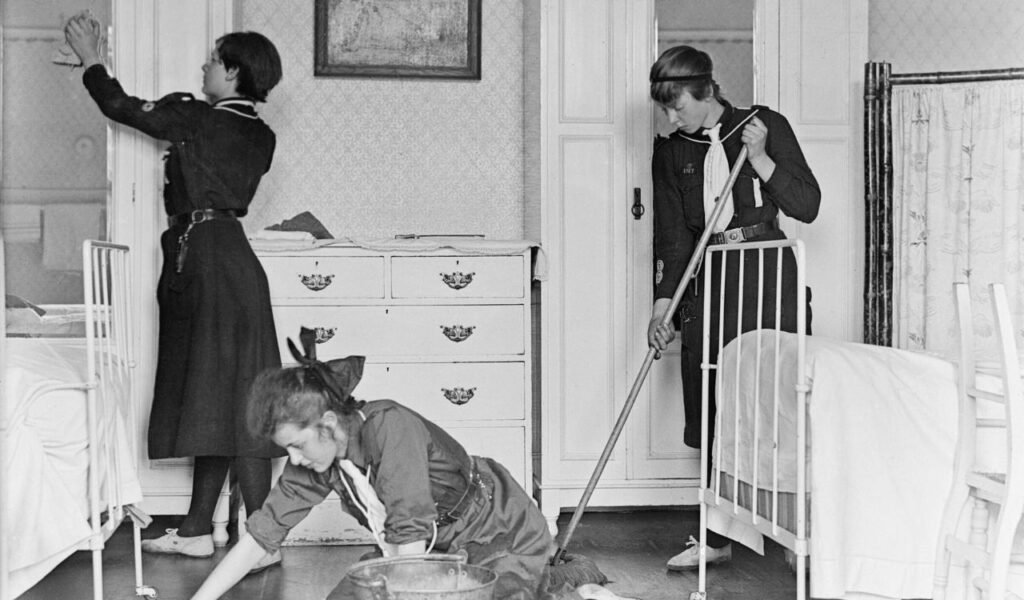 Three women in vintage uniforms clean a small guestroom—one scrubs the floor, another sweeps, and the third dusts a door frame.