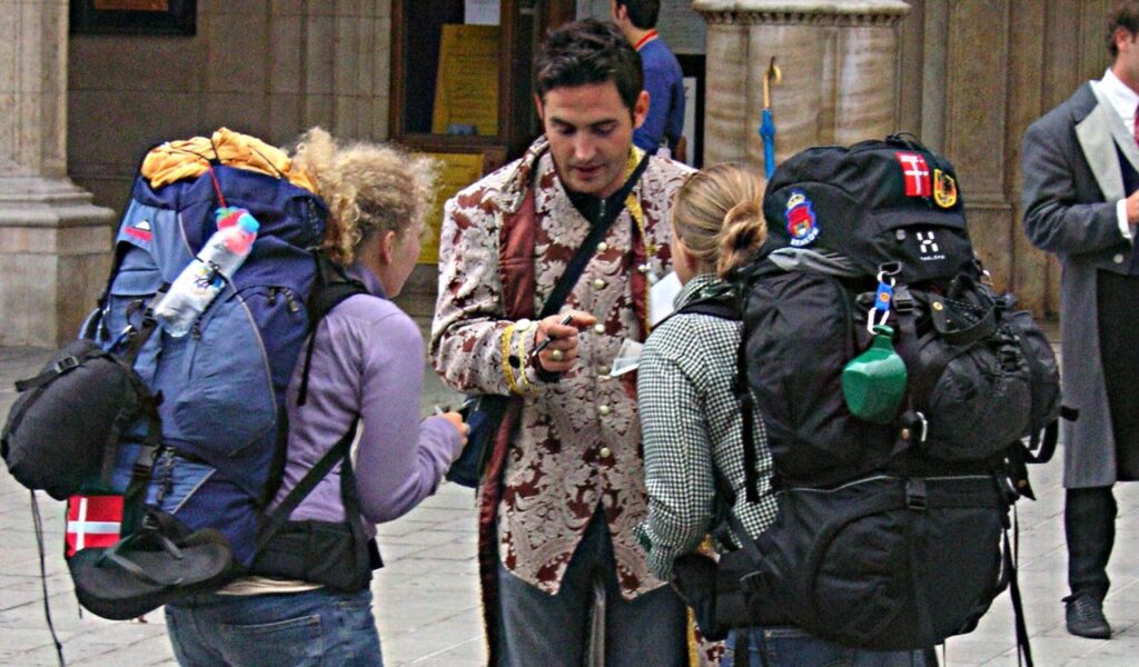 Two backpackers talk to a local guide in ornate clothing on a city street, possibly asking for directions or help.