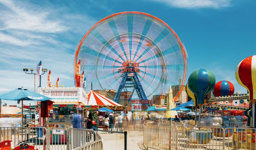 Colorful amusement park scene with a spinning Ferris wheel labeled Wonder Wheel, candy stalls, balloon-shaped rides, and visitors walking under a sunny blue sky.