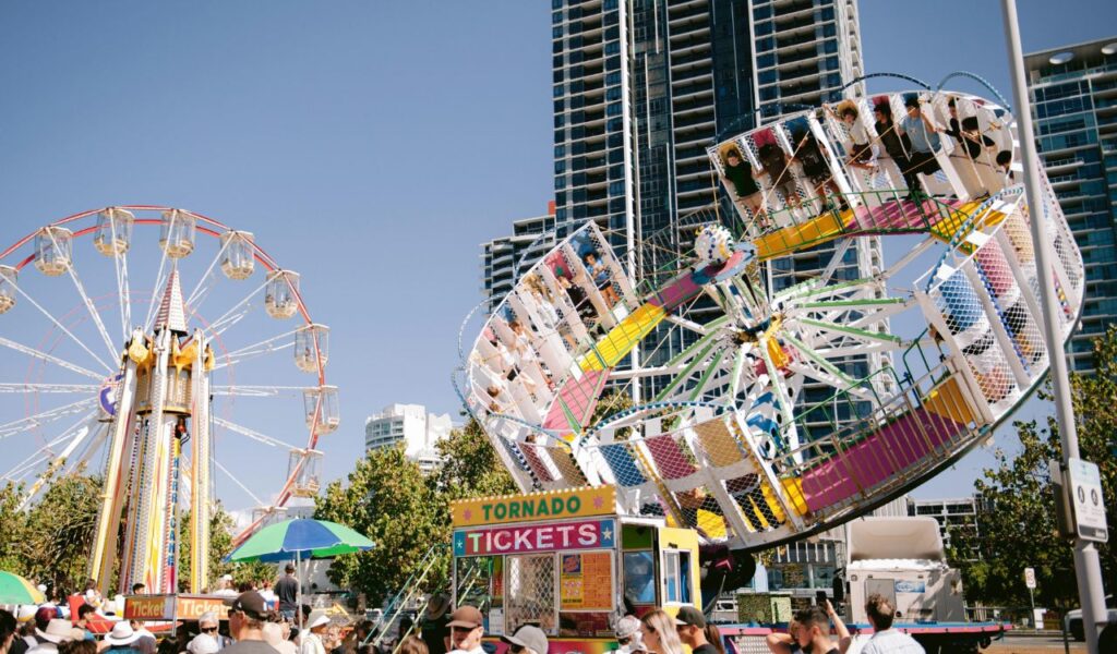 **Alt Text (115 characters):**
Crowds at a vibrant amusement park with spinning rides, a Ferris wheel, and a ticket booth on a sunny day.