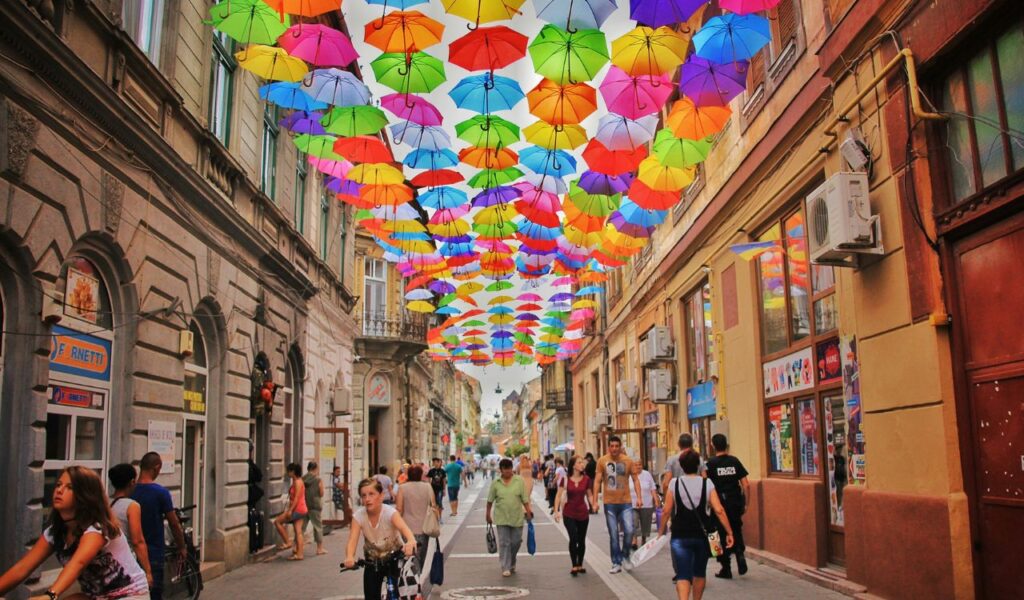 People walk and bike under a colorful canopy of umbrellas suspended above a lively pedestrian street in a historic district.