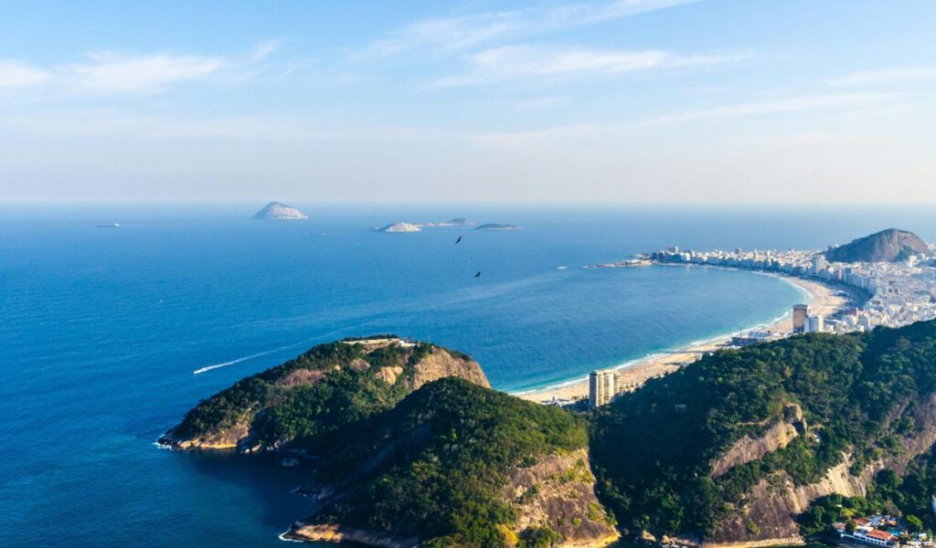 A scenic aerial view of a lush coastal landscape with forested hills, a curved beach, and distant islands across a calm blue ocean.