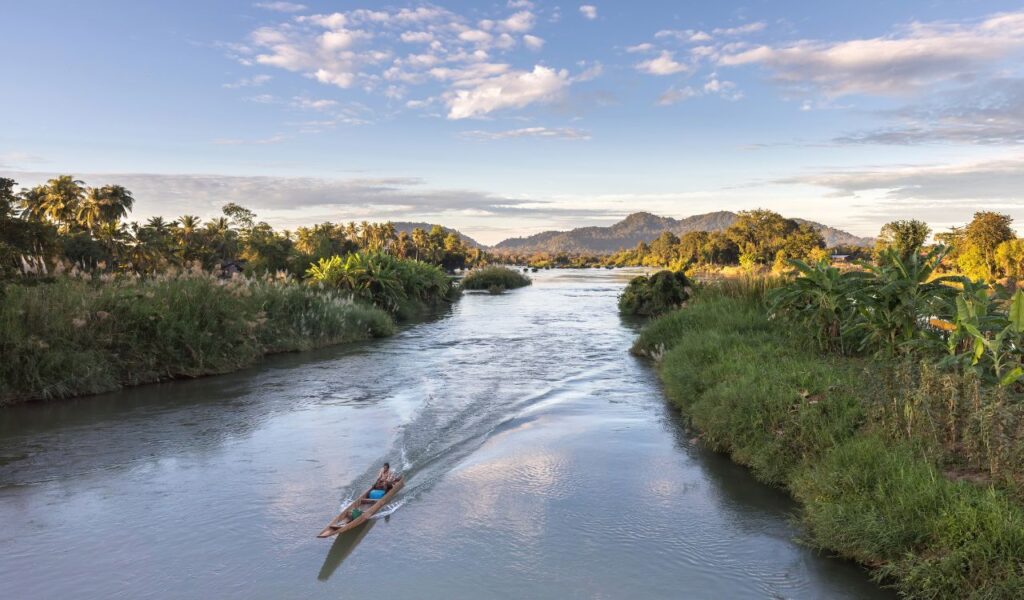 Small wooden boat traveling on a narrow, low-level river surrounded by lush greenery and distant hills at sunset.