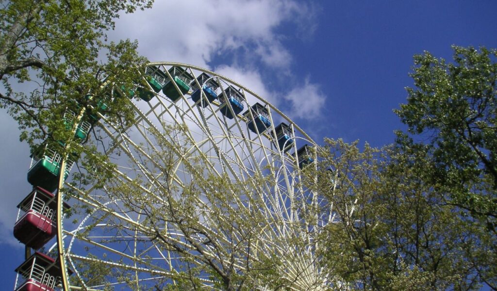 Giant Ferris wheel framed by green trees and blue sky, capturing a fun outdoor adventure scene on a sunny day.