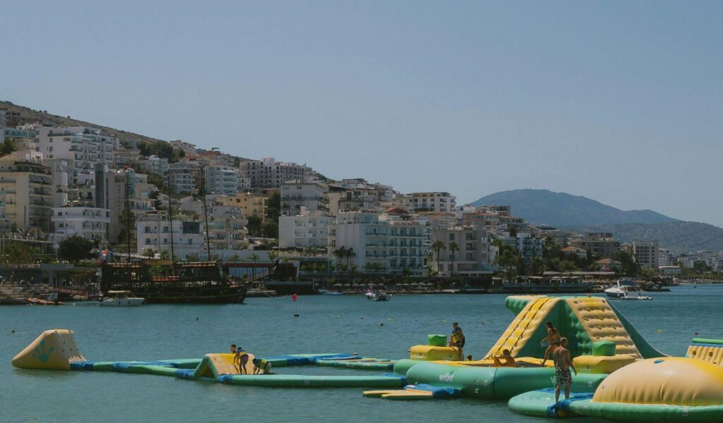 People play on inflatable water structures in the sea near the Albanian Riviera, with hillside buildings and mountains in the background.