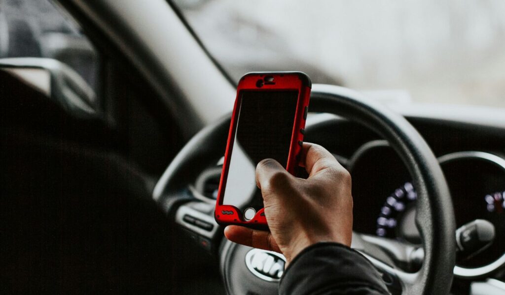 A driver holds a smartphone in one hand while sitting behind the wheel of a car, indicating distracted driving and potential phone use while in motion