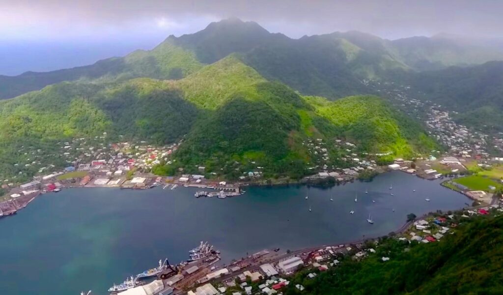 Aerial view of a harbor town in American Samoa surrounded by lush green mountains under a cloudy tropical sky.