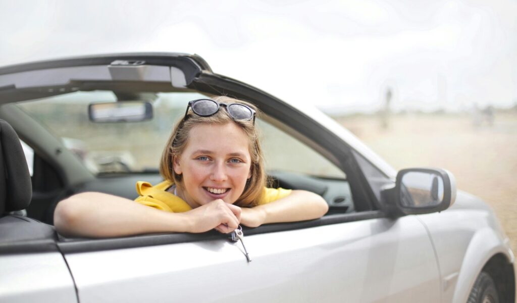 Smiling young woman leaning out of a convertible car window, enjoying a carefree road trip moment on a bright day.