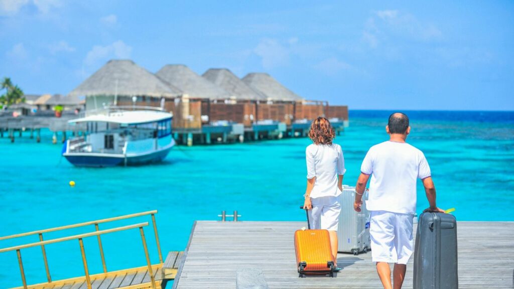 A couple walks along a wooden pier over turquoise water, pulling suitcases toward overwater villas at a tropical island resort with a boat docked nearby.