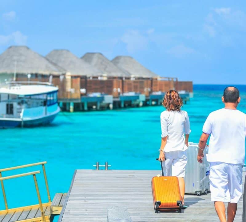 A couple walks along a wooden pier over turquoise water, pulling suitcases toward overwater villas at a tropical island resort with a boat docked nearby.