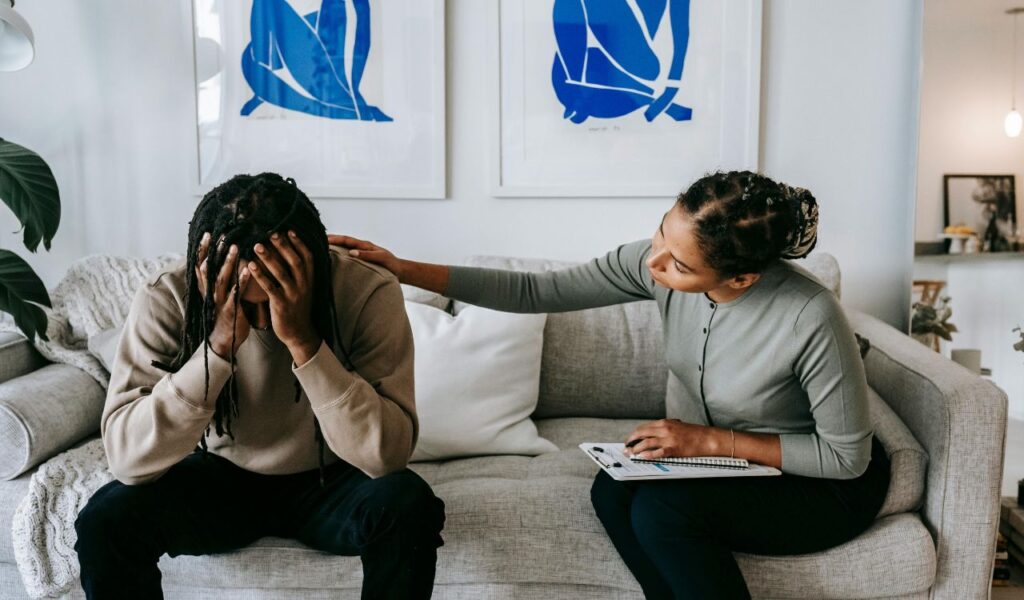 Woman comforting a distressed man on a couch, offering emotional support with a hand on his shoulder.