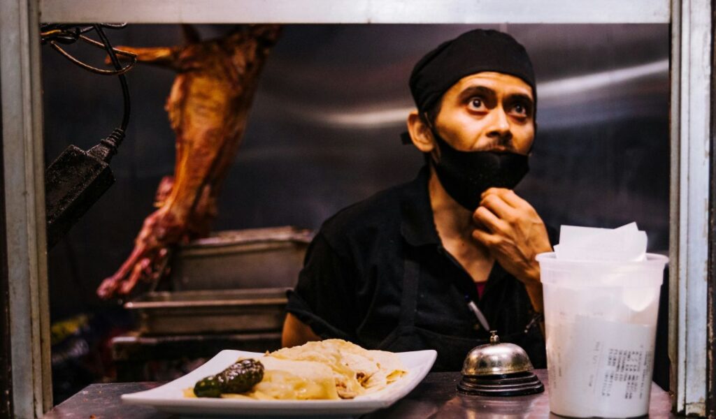 A local street food vendor in a black cap and apron prepares tacos behind a food stall, with a plate of food and receipts in front of him.