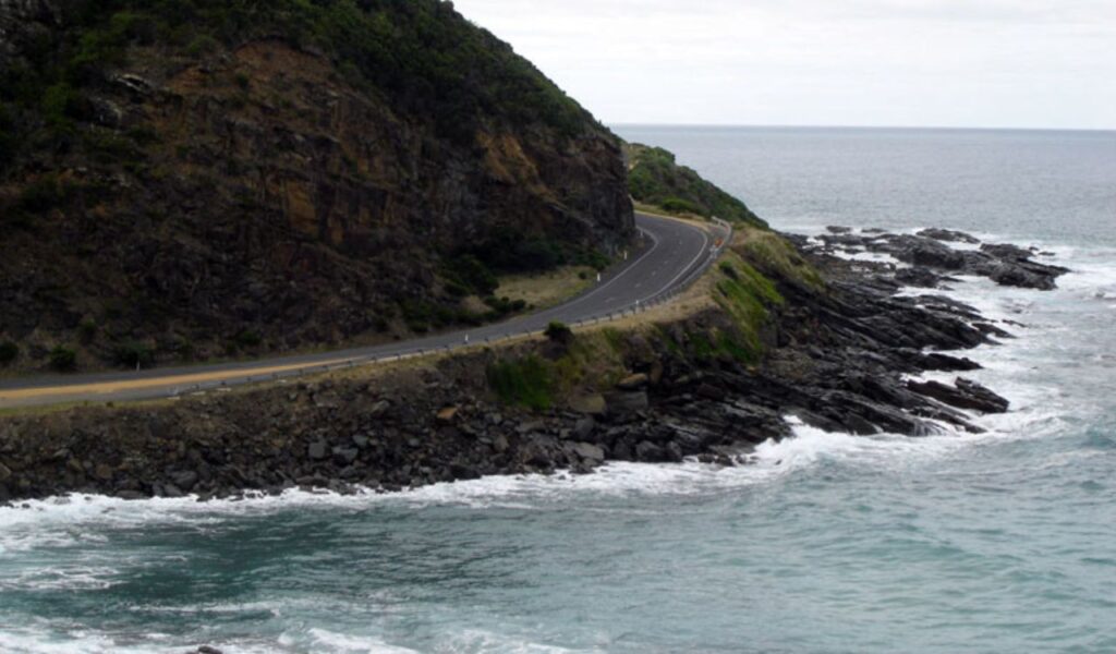 Winding stretch of Australia's Great Ocean Road hugging a rocky coastline with waves crashing below.